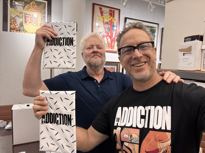 Two men smile for a photo, with one holding up a black and white speckled book titled 'ADDICTION'.