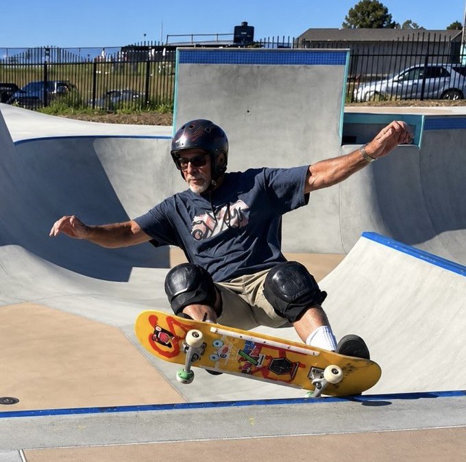 A gray-bearded man wearing a black helmet, sunglasses, knee pads, and a dark "Pepsi" t-shirt performs a skateboarding trick on a ramp at Malibu Bluffs skatepark, with his sticker-covered deck tilted mid-air. The image visually captures the post's claim of a 65-year-old PulseChain advocate enjoying an active, outdoor lifestyle while promoting the cryptocurrency.