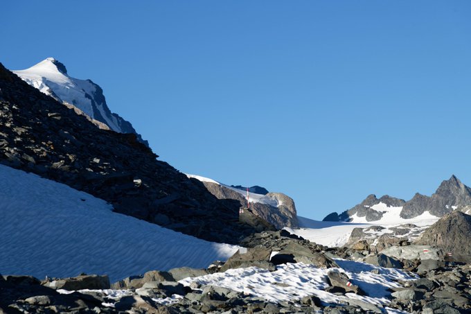 A rugged, rocky mountain landscape is partially covered in snow and glaciers under a clear blue sky, with a small flag visible on a distant ridge.