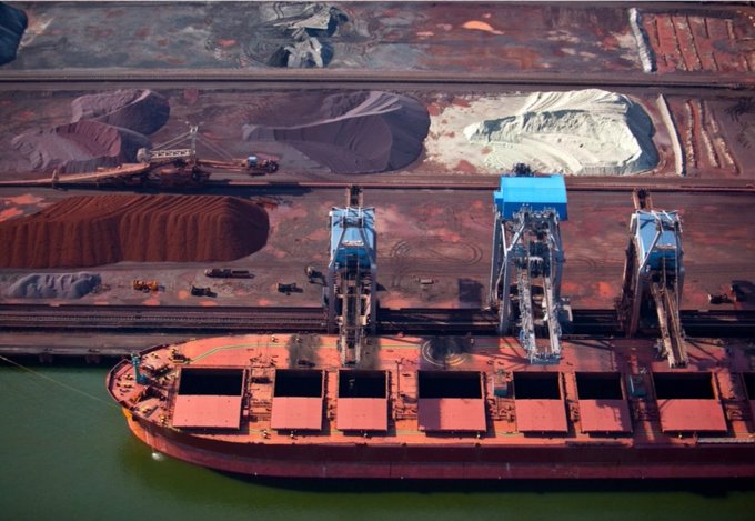 An aerial view of a large red cargo ship docked at a port with blue cranes loading bulk materials.