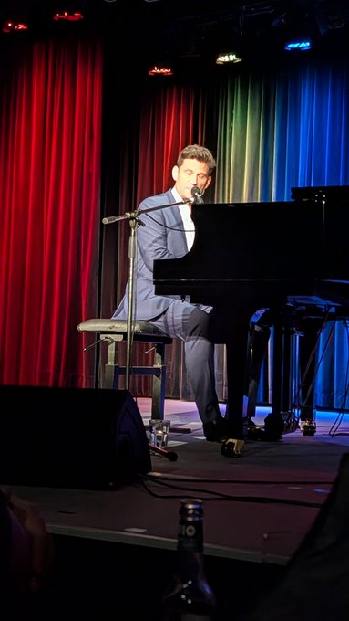 A man in a blue suit sits at a black grand piano on a stage with red and blue curtains, singing into a microphone.