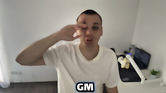 A man in a white t-shirt sits indoors making a salute/pointing gesture toward the camera with a surprised expression, with the letters 'GM' overlaid at the bottom of the frame.
