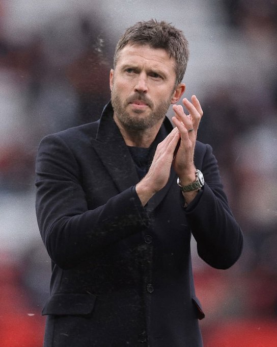 A man with short brown hair and a beard, wearing a dark coat, claps his hands while wearing a watch, appearing to be at a sporting event with blurred red stadium seating in the background.