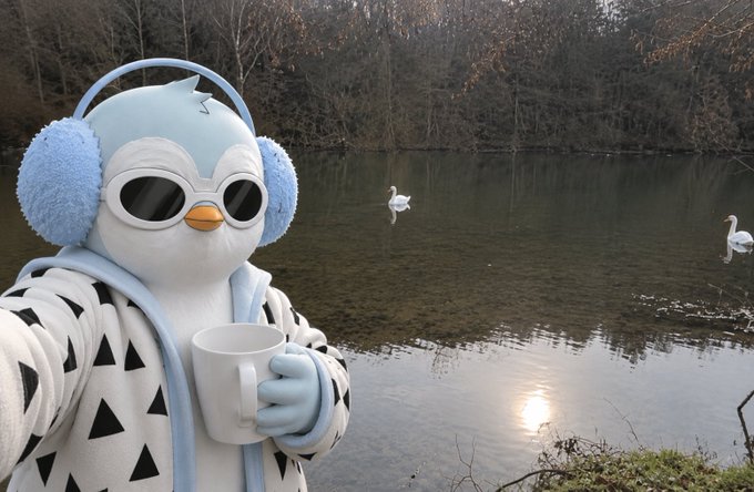 A cartoon penguin character wearing blue earmuff headphones, sunglasses, and a black and white patterned hoodie holds a white mug by a lake with two swans swimming in the background.
