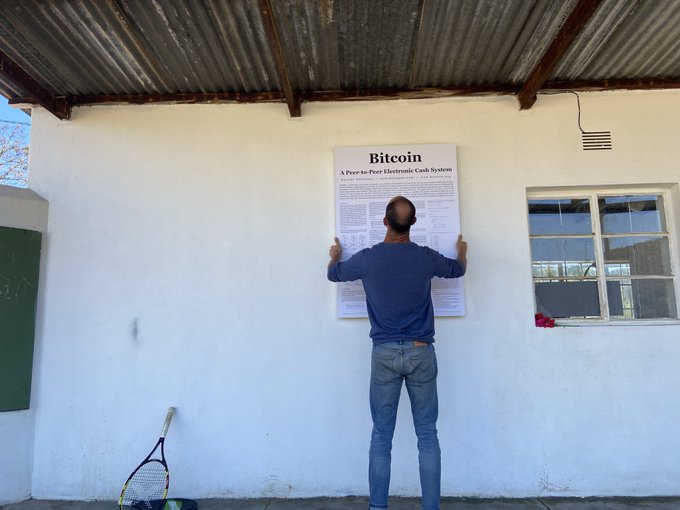 A man stands with his back to the camera, mounting a large poster titled 'Bitcoin: A Peer-to-Peer Electronic Cash System' on a white exterior wall under a corrugated metal roof.