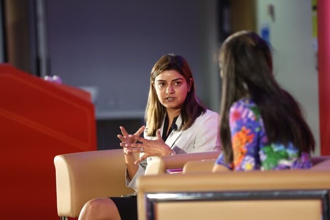 The image shows two women sitting next to each other, engaged in conversation. One woman appears to be wearing a white blouse while the other has a colorful floral top. They are both seated on chairs placed around a table. The background features a red wall with a sign that reads "Ripple's own researcher just said the quiet part out loud anonymous transfers on a public blockchain that' s a harder problem to solve". This suggests that they might be discussing recent developments in cryptocurrency or blockchain technology, as Ripple is known for its contributions to this field.