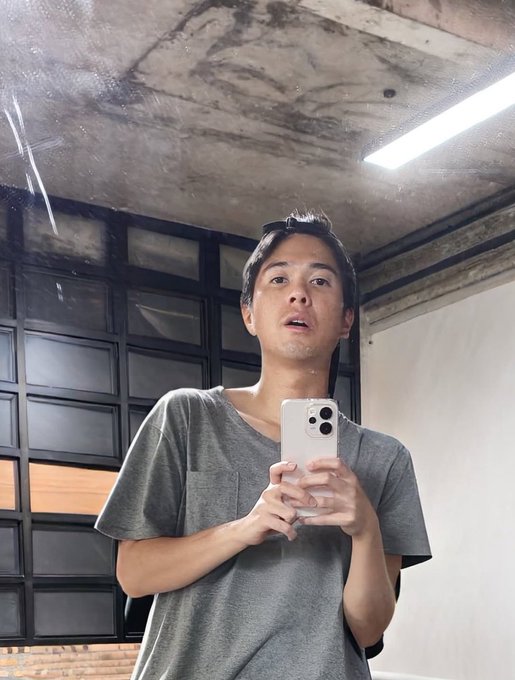 A man in a gray t-shirt taking a mirror selfie with a silver smartphone in an industrial-style room with a concrete ceiling and a grid-patterned window.