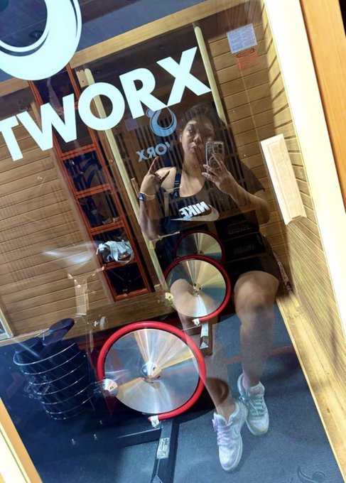 A woman taking a mirror selfie in a gym with a 'WORX' sign, wearing a Nike sports bra and shorts, standing near weight plates on a machine.
