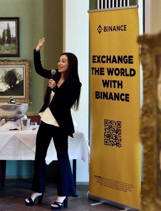 A woman in a black suit waving while holding a microphone next to a yellow 'BINANCE - Exchange the World with Binance' banner featuring a QR code.