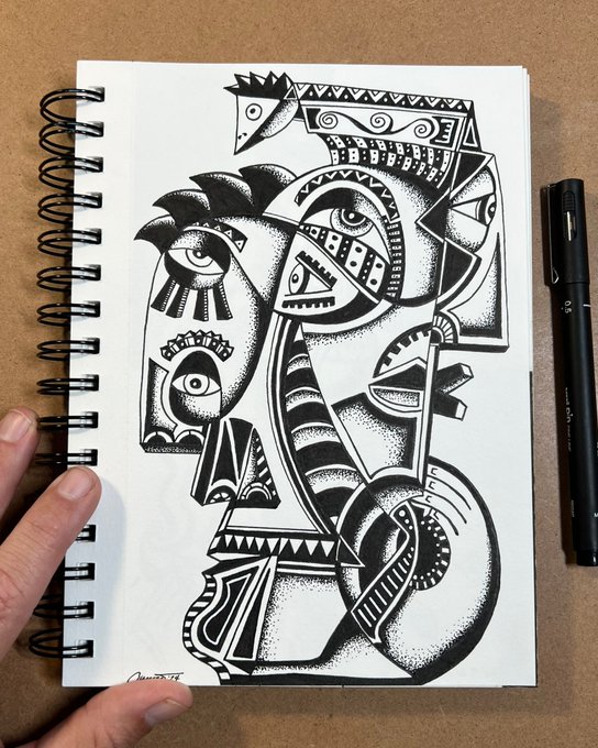 A black and white stippled ink drawing in a spiral notebook depicts an abstract tribal-style figure with geometric patterns and eyes, with a black marker pen resting beside it on a brown surface.