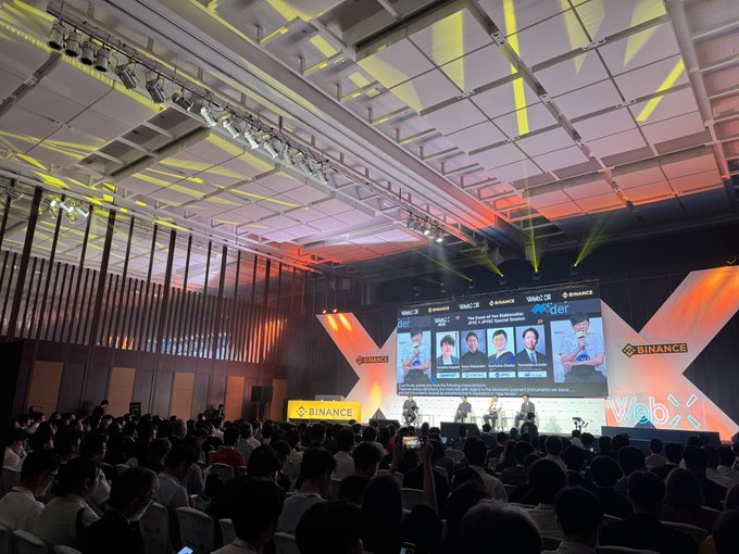 A large conference audience gathers at the Binance WebX event, watching a panel discussion on a big screen featuring the "JPYC × JPYSC" session about The Bank of Japan's stance on tokenization, with the main stage and branded Binance backdrop brightly lit on Day 2 of the conference.