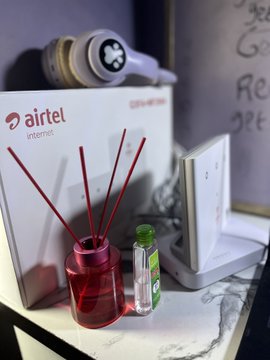 A small tabletop scene featuring an Airtel Internet branded box, a reed diffuser with red sticks, and a small green bottle. The phrase "e no mean" connects to the image's casual, everyday Nigerian workspace vibe, suggesting the setup—complete with visible "airtel internet" text—is humble yet meaningful to the poster.