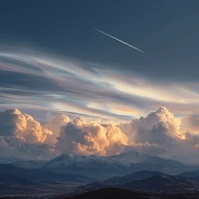 A scenic view shows a jet contrail streaking across a twilight sky with dramatic clouds above snow-capped mountains.
