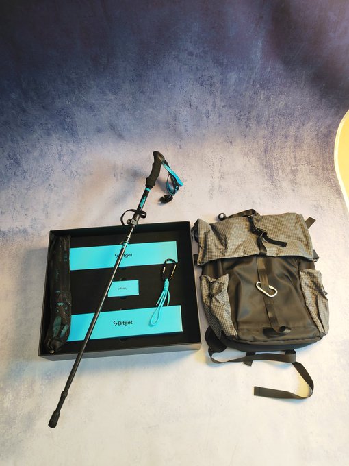 A Bitget-branded box containing a trekking pole with blue lanyard, displayed next to a gray/green tactical-style backpack with multiple compartments and a carabiner.
