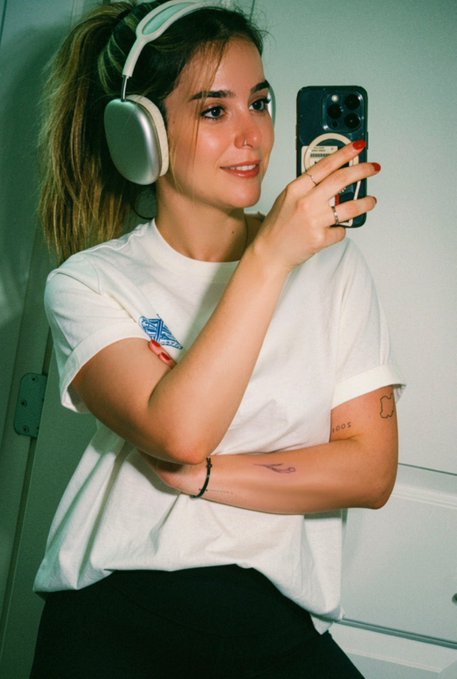 A young woman with long hair wears silver headphones and a white t-shirt while taking a mirror selfie with her smartphone.