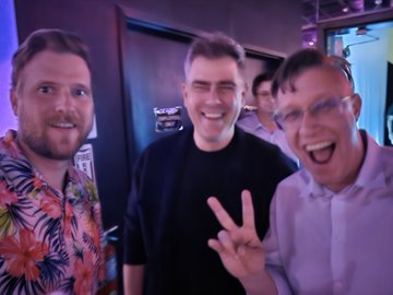 Three men posing together for a photo in a dimly lit venue; the man on the right wears glasses and makes a peace sign, while the center man smiles broadly in a black shirt.