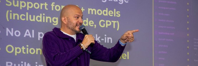 A bald man in a purple hoodie speaks into a microphone while pointing, with a presentation slide behind him mentioning 'Support for AI models (including Claude, GPT)' and 'No AI p*** k-in Option'.