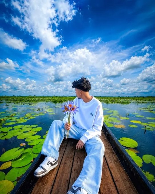 A young man in a white sweatshirt and light jeans sits on a wooden boat, holding purple lotus flowers while floating among lily pads under a bright blue cloudy sky.