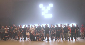 A group photo of a large team of people posing together in a dark room, illuminated by bright stage lights arranged in a Y-shaped configuration above them.