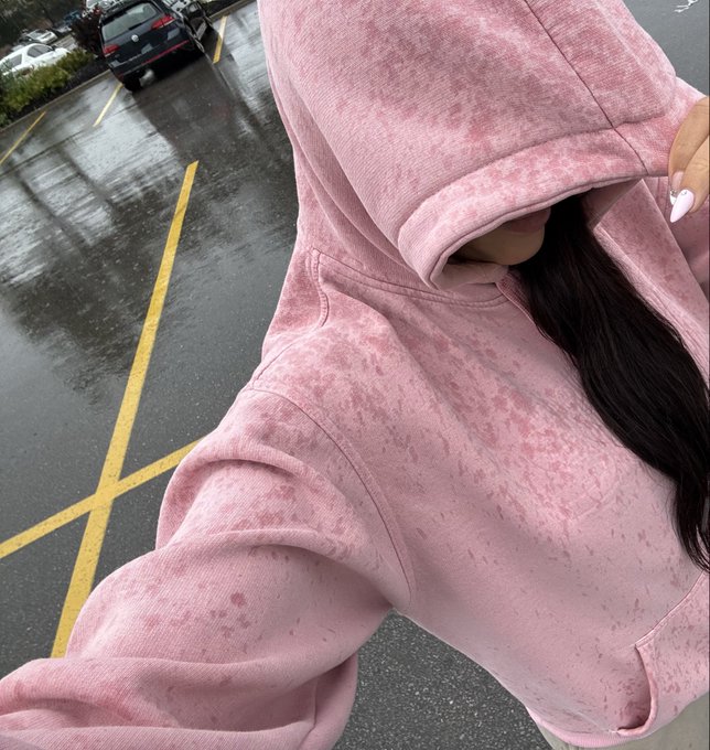 A person wearing a pink acid-wash hoodie pulled up to cover most of their face, with long dark hair flowing out, standing in a rainy parking lot with yellow lines visible on the wet pavement.