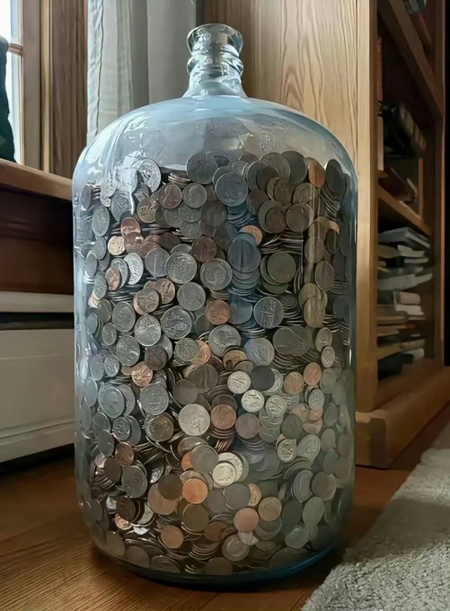 A large clear glass jug or bottle filled with a massive collection of mixed silver and copper coins sitting on a wooden floor.