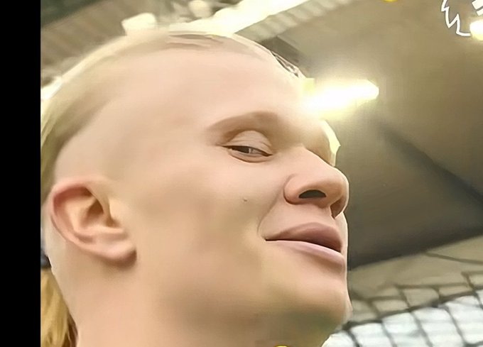 A close-up of a blonde-haired humanoid robot with a smiling face displayed in a brightly lit indoor setting.