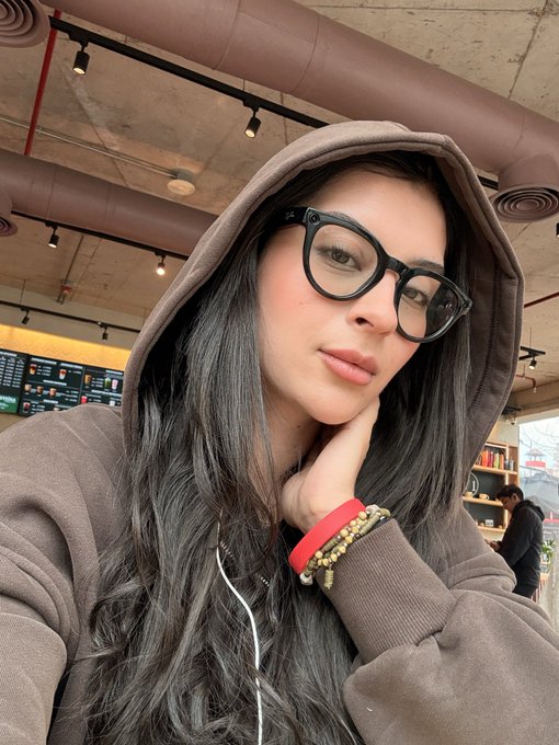 A selfie of a young woman with long dark hair wearing black-rimmed glasses and a brown hooded sweatshirt, resting her chin on her hand.