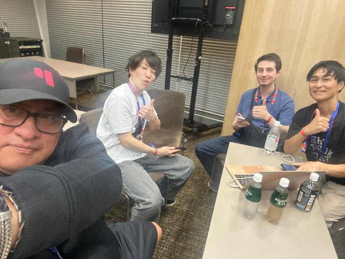 Four men wearing lanyards pose for a selfie in an indoor lounge, with two giving thumbs up and beverages on the table.