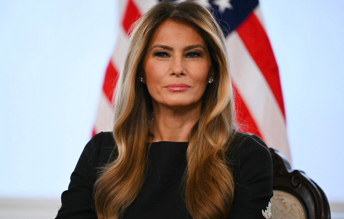 The image features a woman with blonde hair sitting in front of an American flag, looking directly at the camera. The background behind her appears to be white, which contrasts nicely with her blonde hair. The overall scene suggests that she might be participating in an event or gathering related to the United States, such as a political speech or a patriotic celebration.