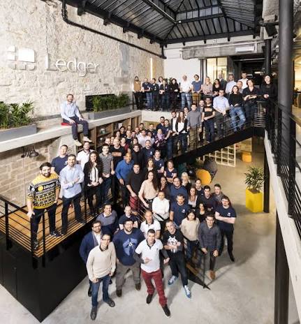 A large group photo of employees gathered in a modern, industrial-style office space with a sign reading 'Ledger' on a stone wall.