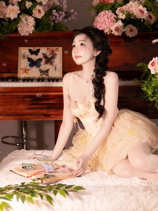 A young woman in a pale yellow floral dress kneels by a piano, surrounded by pink flowers and a butterfly display, holding an open book.