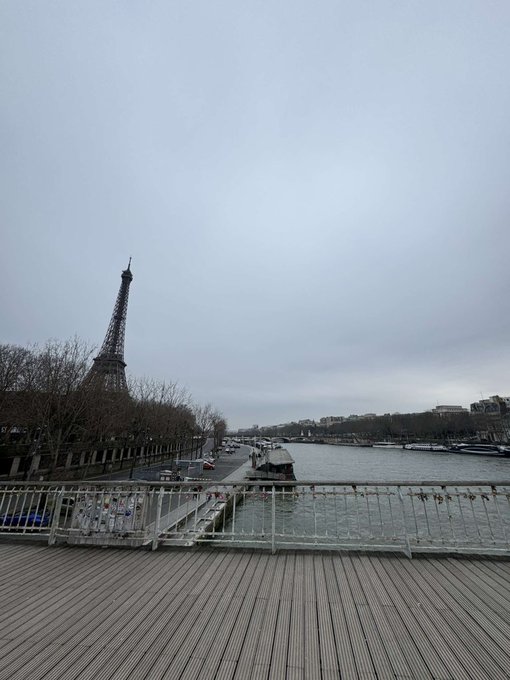 An overcast view of the Eiffel Tower from across the Seine, taken from a wooden deck.