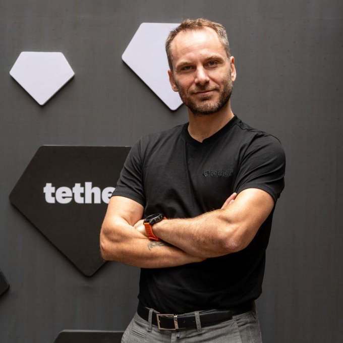 A man wearing a black "tether" t-shirt stands with arms crossed in front of a branded wall featuring hexagonal logos and a "tether" sign, fitting a story about the company's Tether token and its exposure to struggling offshore bank EQIBank.