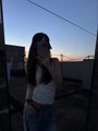 A blurry low-light photo of a woman with long dark hair standing outdoors at dusk, possibly on a rooftop, wearing a white top and blue jeans.