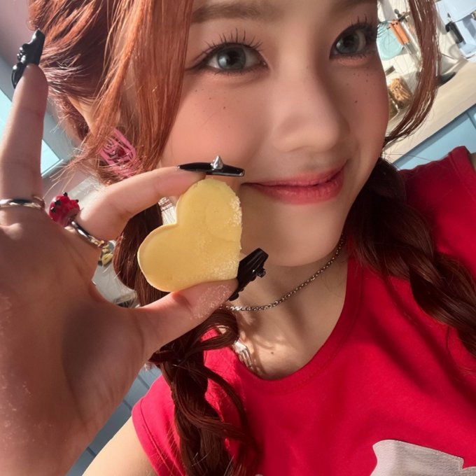 A young woman with reddish-brown hair, styled long nails, and pink accessories holds up a heart-shaped cookie dusted with sugar to the camera.