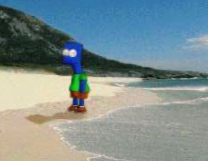 A blue cartoon character wearing green shorts and a green shirt stands on a sandy beach near the water.