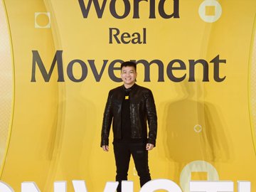 A man in a black leather jacket stands in front of a bright yellow backdrop displaying the text 'World Real Movement' and 'INVEST'.