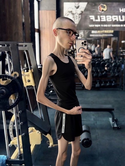 A mirror selfie of a slim, shaved-head person wearing glasses, a black tank top, and black shorts, standing in a gym with weight equipment visible behind them.