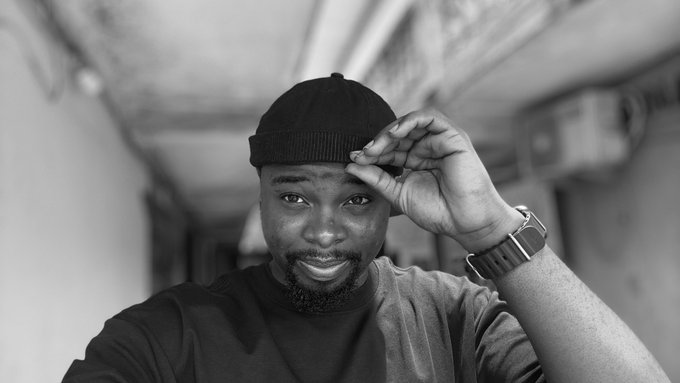 A black and white selfie shows a smiling man with a beard wearing a dark t-shirt and a beanie, with an Apple Watch visible on his raised wrist.