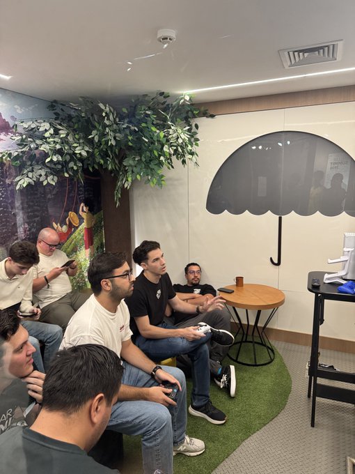 A group of men sit on tiered seating playing video games in a casual lounge area decorated with a large artificial tree, a grass patch, and an umbrella decal, capturing a relaxed break at the Build On Stellar Builder Summit in São Paulo.