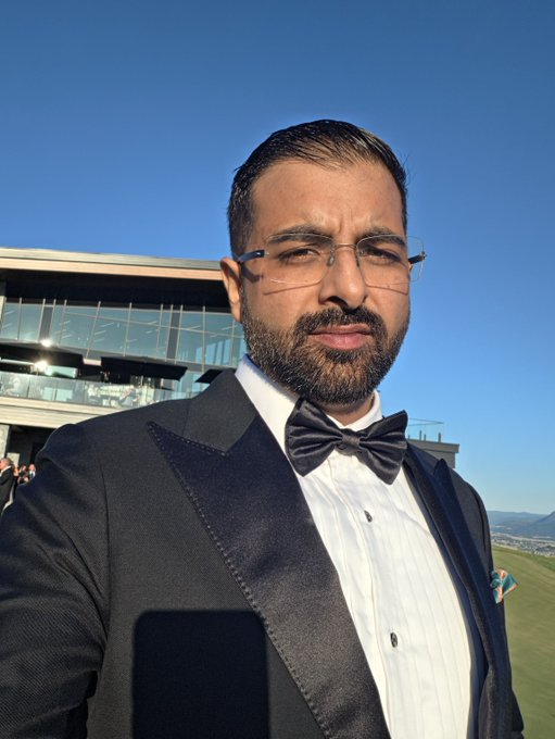 A man with a beard and glasses, wearing a black tuxedo with a white shirt, black bow tie, and pocket square, takes a selfie outdoors in front of a modern building and green field.