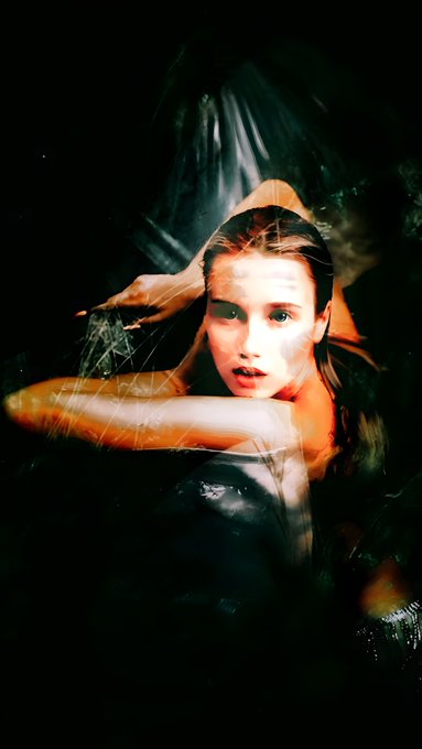 A moody, surreal photo of a young woman with wet hair looking directly at the camera, her arm raised across her face against a dark, shadowy background.