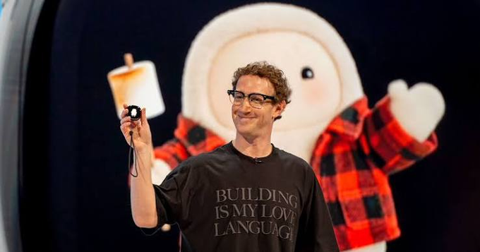 A curly-haired man wearing glasses and a black "BUILDING IS MY LOVE LANGUAGE" t-shirt smiles while holding up a small camera, with a large mascot character resembling a marshmallow in a red plaid jacket standing behind him on stage. The image appears to relate to a playful "$META" reference, with the on-stage product reveal moment suggesting a tech company keynote unveiling.