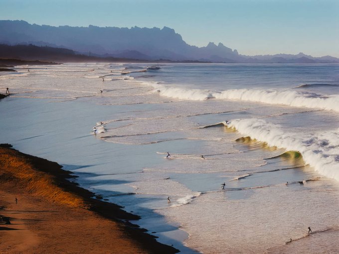 A sweeping coastal scene captures surfers gliding along rolling waves beneath a jagged mountain range, its serene shoreline bathed in golden light that evokes the tranquil escape of a beachside resort. The composition mirrors the post's reference to a "Beachside Resort" by immersing viewers in the natural beauty and leisurely atmosphere of a coastal retreat.