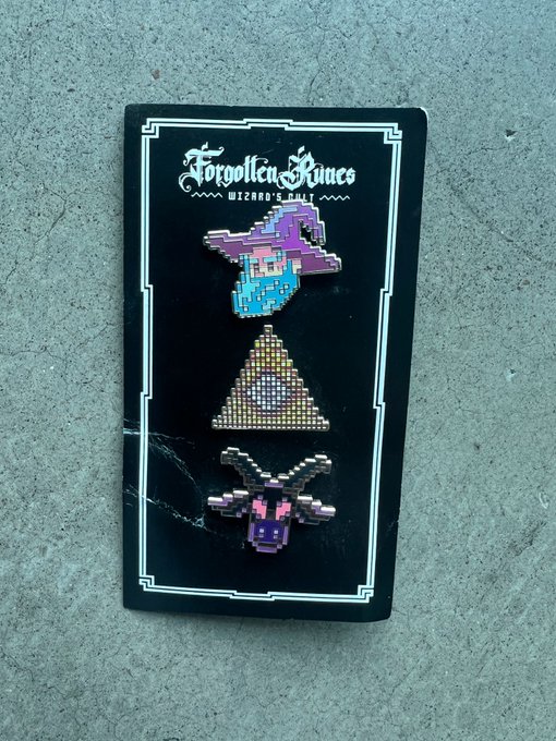A black backing card labeled 'Forgotten Runes Wizard's Cult' displaying three pixel-art style enamel pins featuring a wizard hat, a triangle, and a skull design.