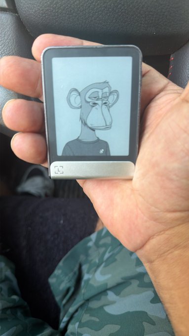 A hand holds a small electronic device displaying a Bored Ape Yacht Club NFT image, with the person wearing camouflage pants and seated in a car.