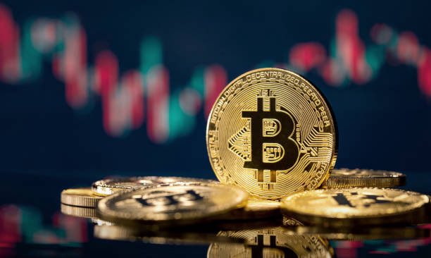 A stack of physical gold Bitcoin tokens with the Bitcoin "₿" logo, set against a background of blurred red and green stock market charts.