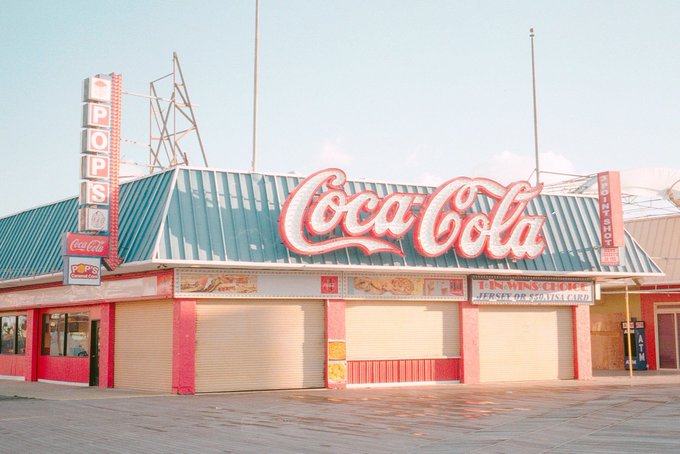 A vintage photograph of a closed beachside Coca-Cola concession stand with a prominent red Coca-Cola sign and a 'POP'S' vertical sign, with Twister and Jersey Girl Souvenirs advertisements visible.