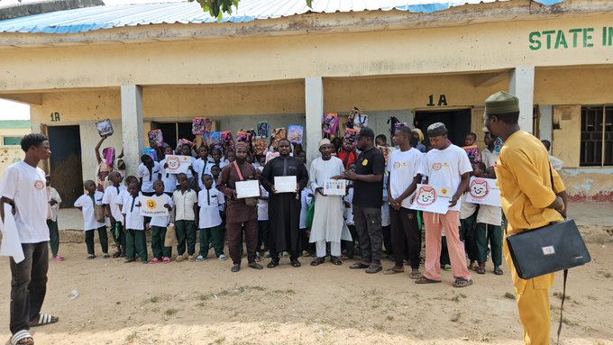 A group of people, including children in white shirts and adults in traditional Nigerian attire, stand outside a building labeled 'STATE II' holding certificates or packages, with a priest and another man in yellow observing.