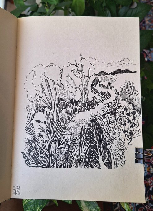 An open sketchbook displays a black ink drawing of a landscape featuring trees, foliage, and mountains, photographed against a backdrop of green plant leaves.
